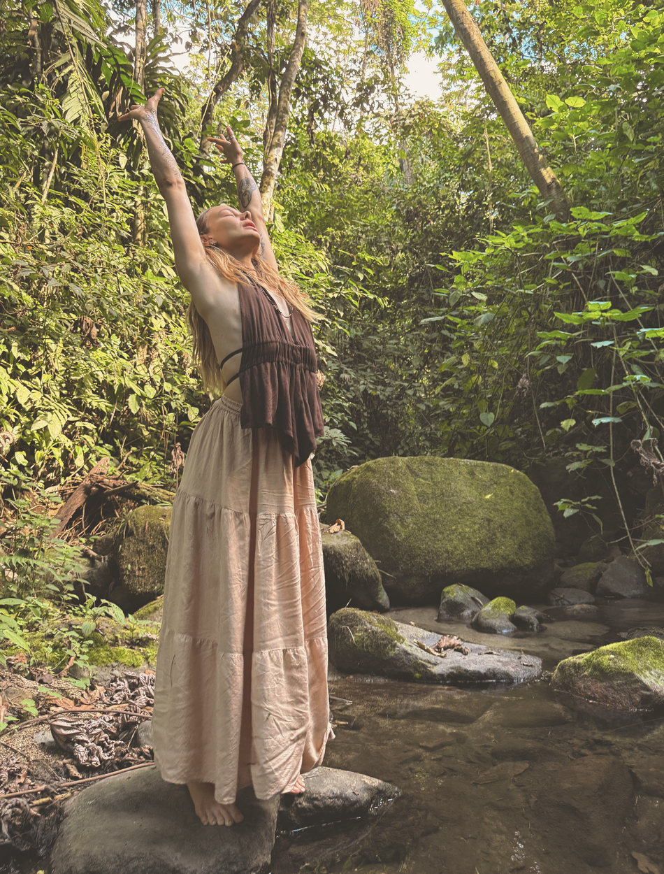 Skye standing barefoot on a rock in the stream, both arms reaching up into the canopy, face lifted.
